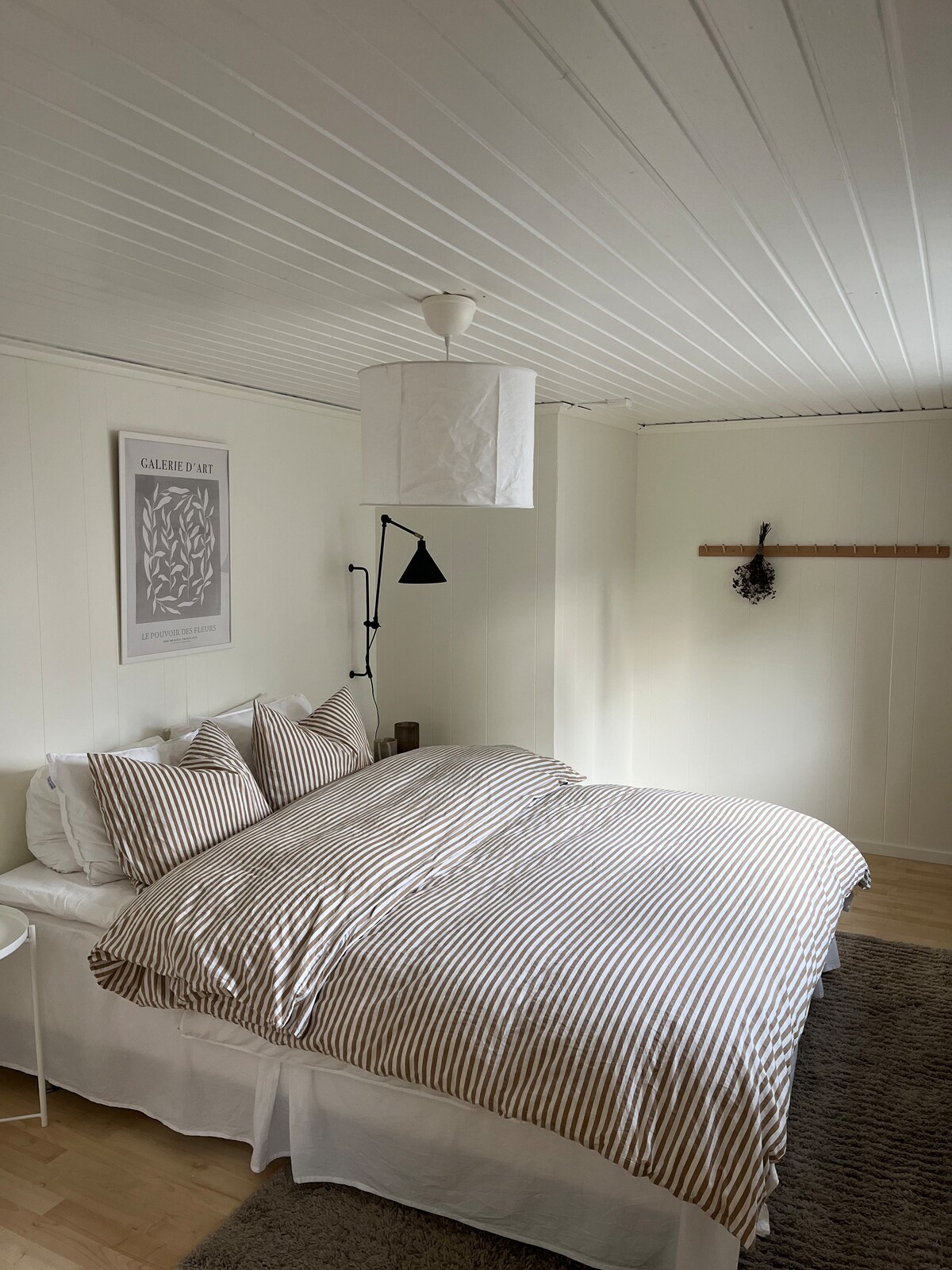 The bedroom features a spacious bed with striped linens arranged neatly. A pendant light hangs above, providing soft illumination. A simple wall art piece is displayed beside a reading light, and a coat rack made of wood adds functional decor. Light-colored walls enhance the airy feel.