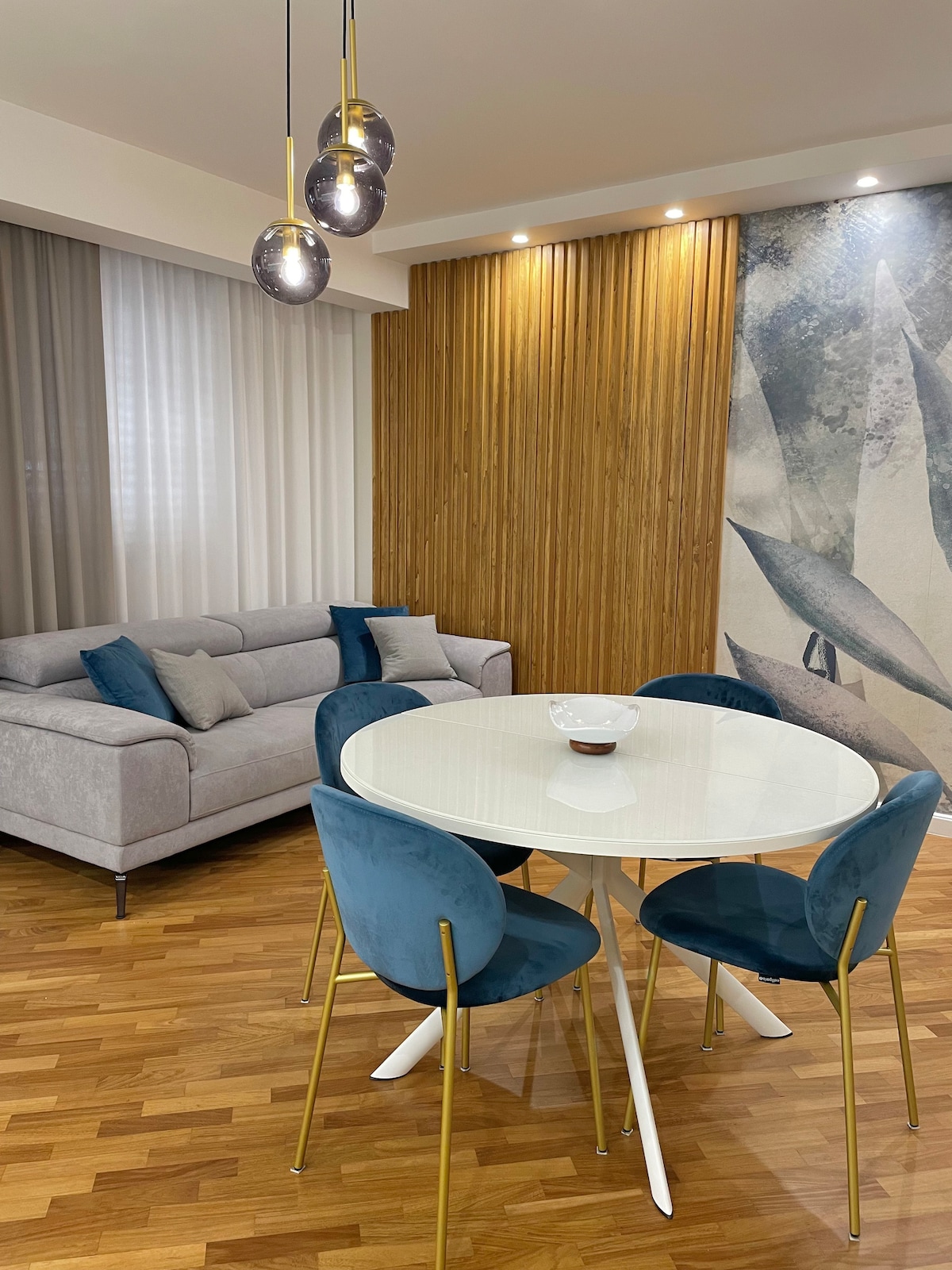 A contemporary dining area is presented with a round white table surrounded by four blue chairs. A modern gray sofa is visible in the background, adorned with decorative cushions. Natural light filters through sheer curtains, softly illuminating the space.