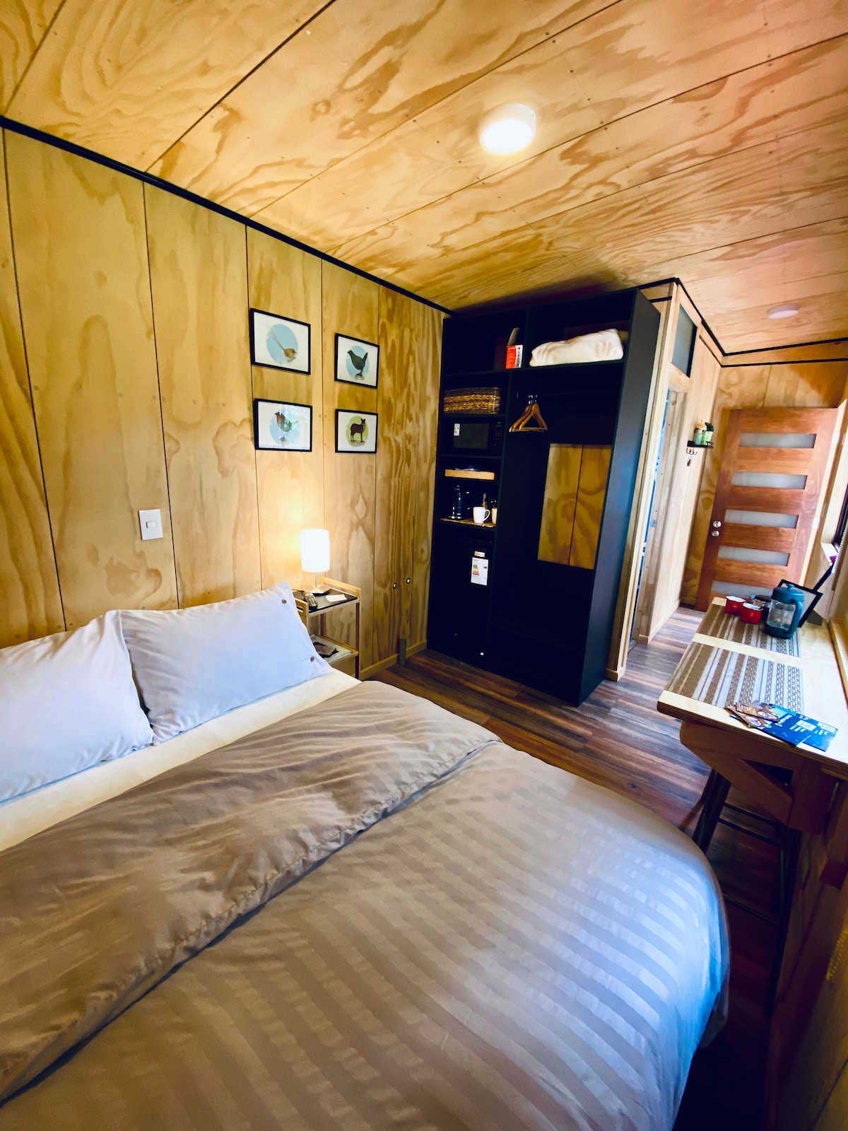 A cozy sleeping area is highlighted by wooden paneling and soft bedding. A small lamp on a bedside table casts a warm glow. Artwork featuring birds is displayed on the wall. A compact kitchenette and a study space are visible, providing practical amenities in a compact layout.