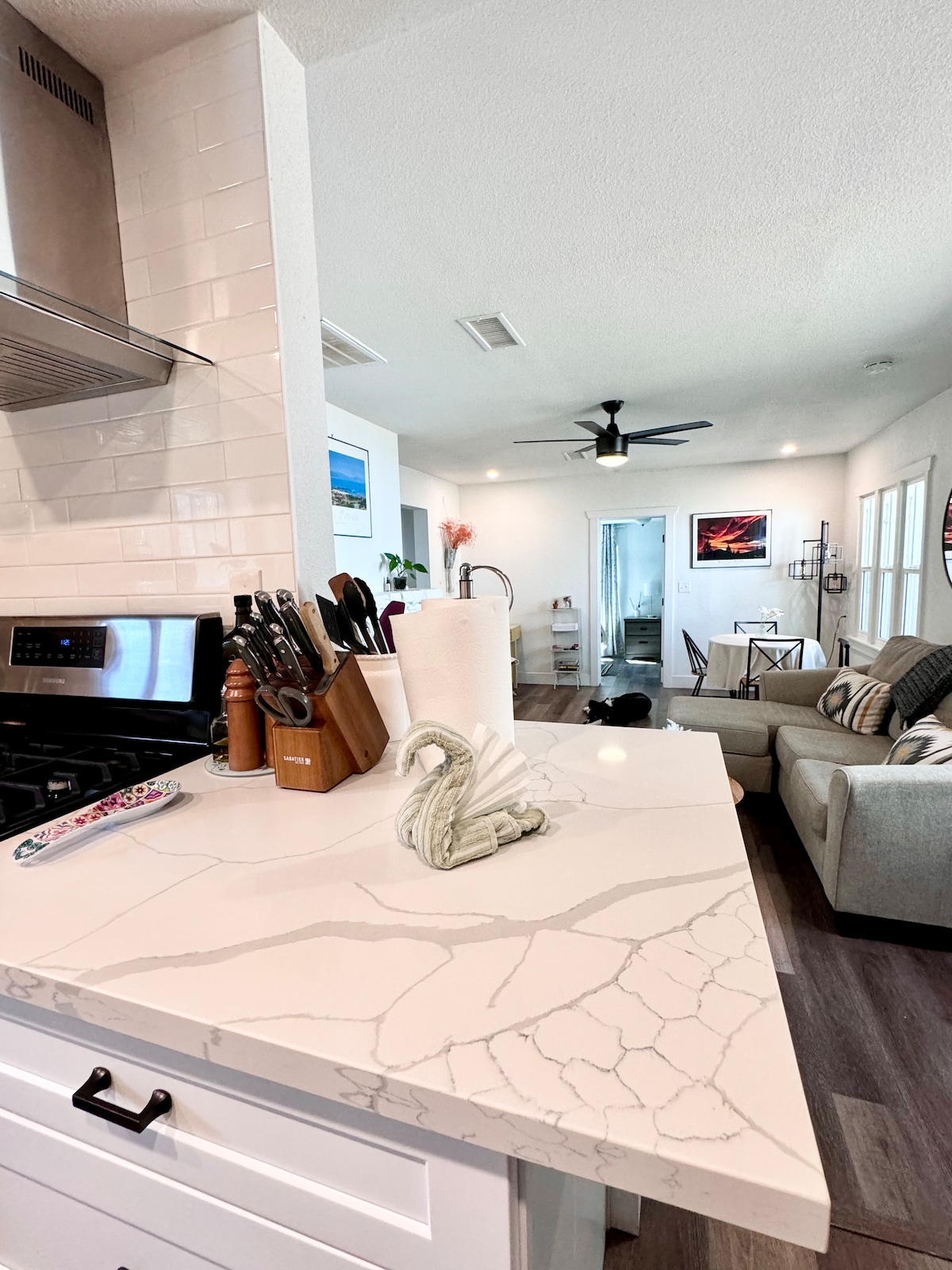 A modern kitchen space is highlighted by sleek white cabinetry and a veined marble countertop. Kitchen utensils are organized neatly in a wooden holder, while a decorative swan figurine adds a subtle touch. The open floor plan flows into a cozy living area, visible in the background.