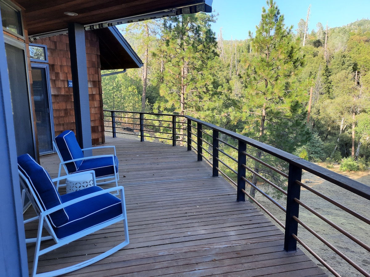 A spacious wooden deck is showcased, featuring two white rocking chairs with blue cushions. The railing provides a clear view of the surrounding trees and landscape, creating an inviting outdoor space for relaxation.