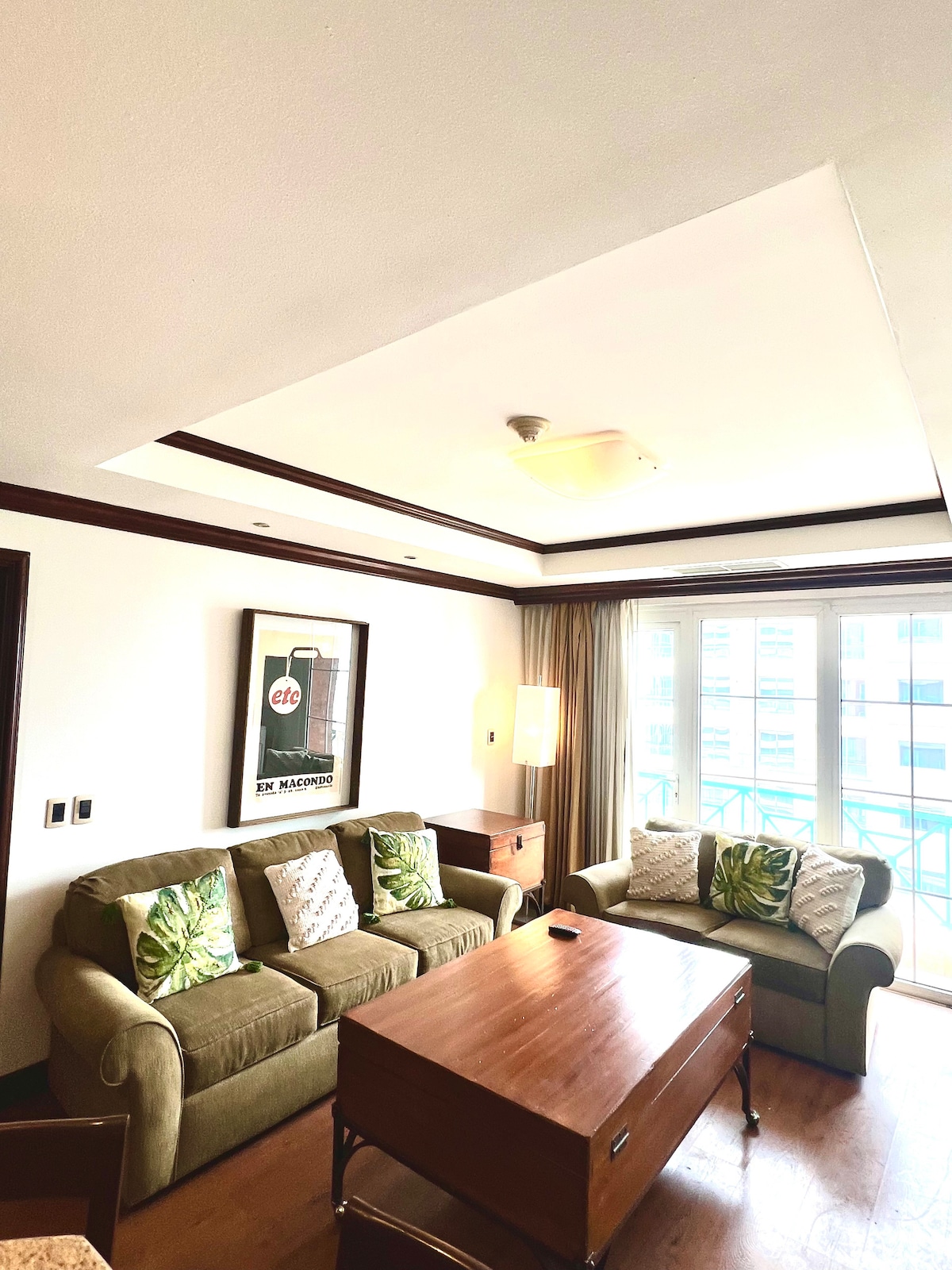 A cozy living area is furnished with a green sofa and a matching armchair, accented by decorative cushions. A wooden coffee table sits in front, with a framed art piece on the wall. Large windows allow natural light to fill the space.