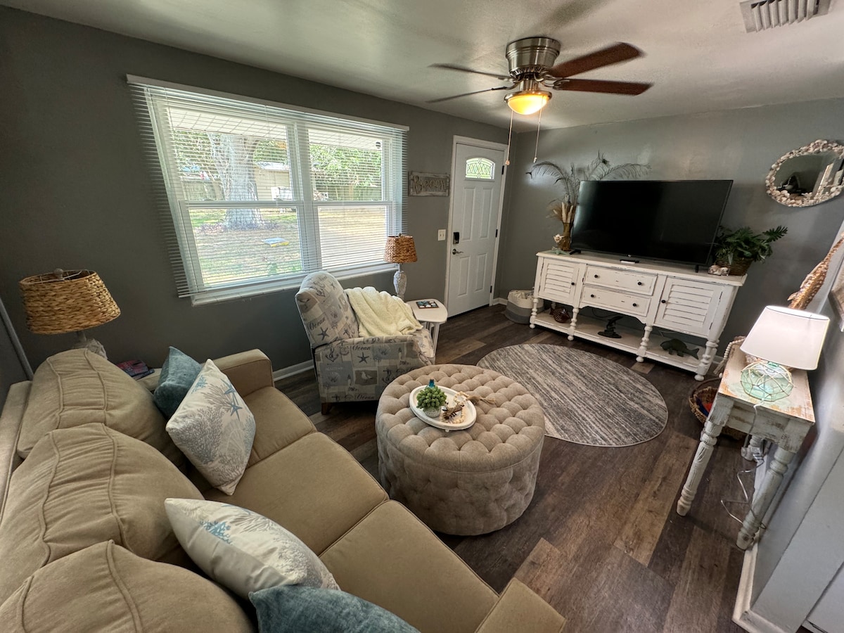 A cozy living area features a light-colored sectional sofa and a plush, tufted ottoman at its center. Natural light floods the space through large windows, complemented by a ceiling fan. A TV is mounted on a white cabinet, while decorative accents enhance the inviting ambiance.