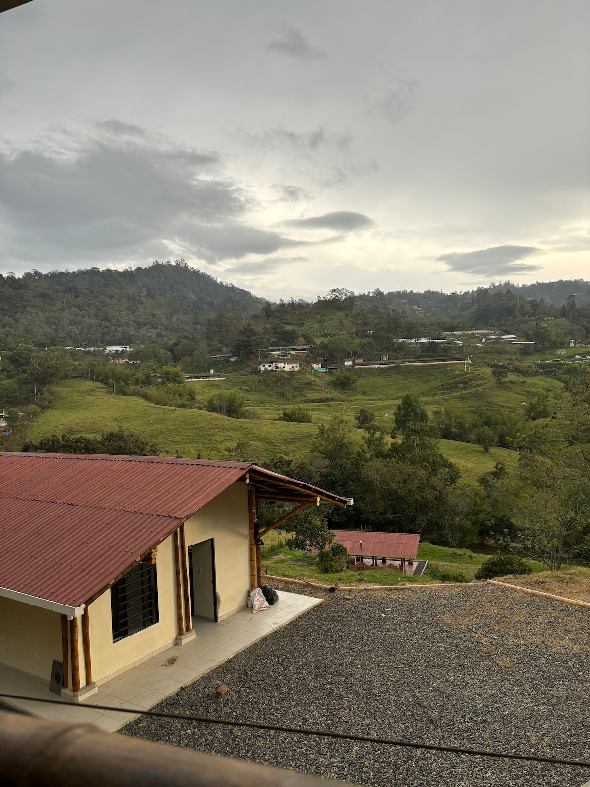 A view of lush, rolling hills is captured, showcasing greenery under an overcast sky. Various structures are visible in the distance, surrounded by trees. A partially visible building with a red roof is framed on the left, providing perspective on the serene landscape.