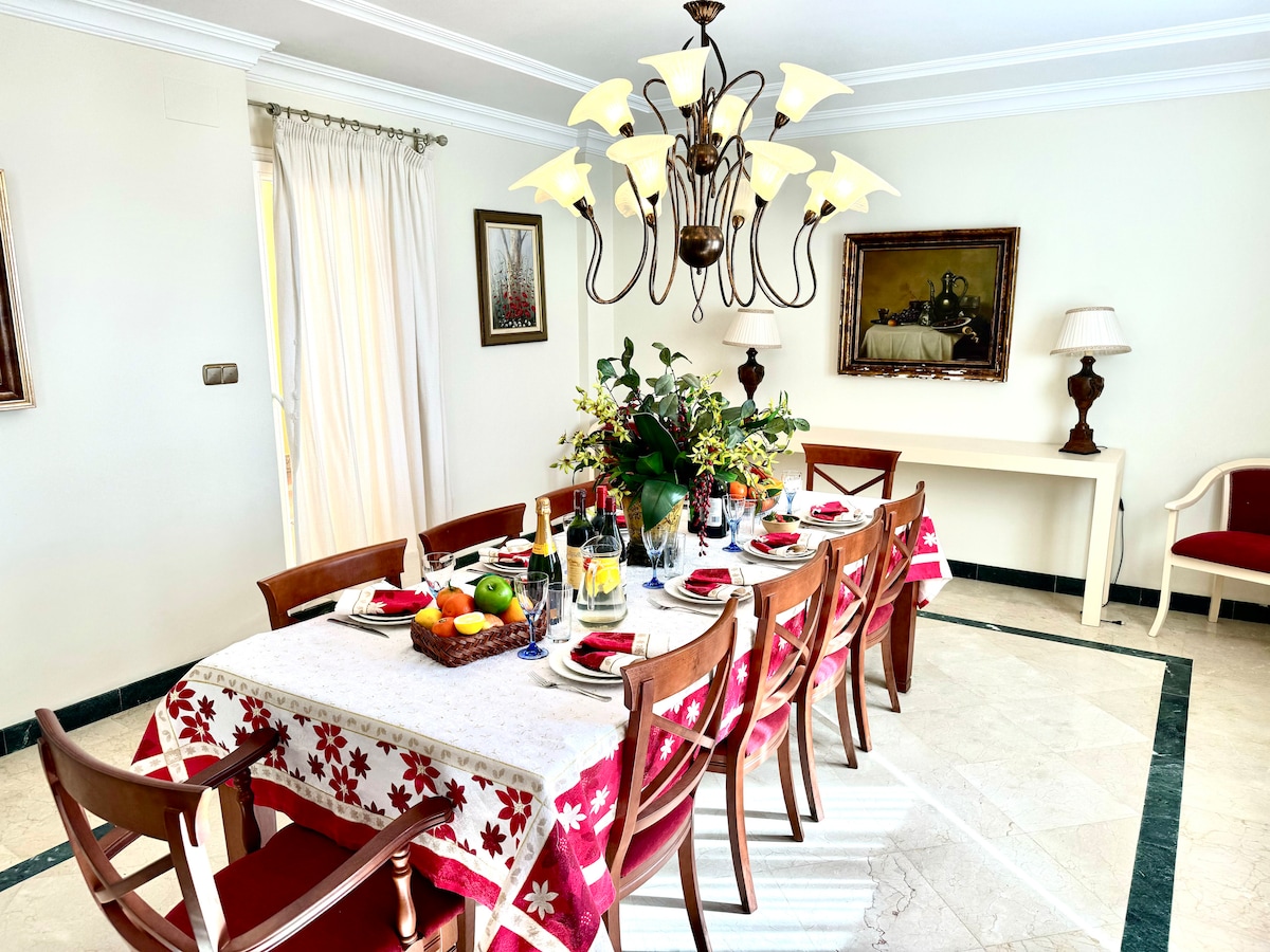 A spacious dining area is highlighted by a large table set for a meal, surrounded by wooden chairs. A chandelier hangs above, complemented by natural light filtering through the sheer curtains. A decorative centerpiece features fresh flowers and fruits, enhancing the inviting atmosphere.
