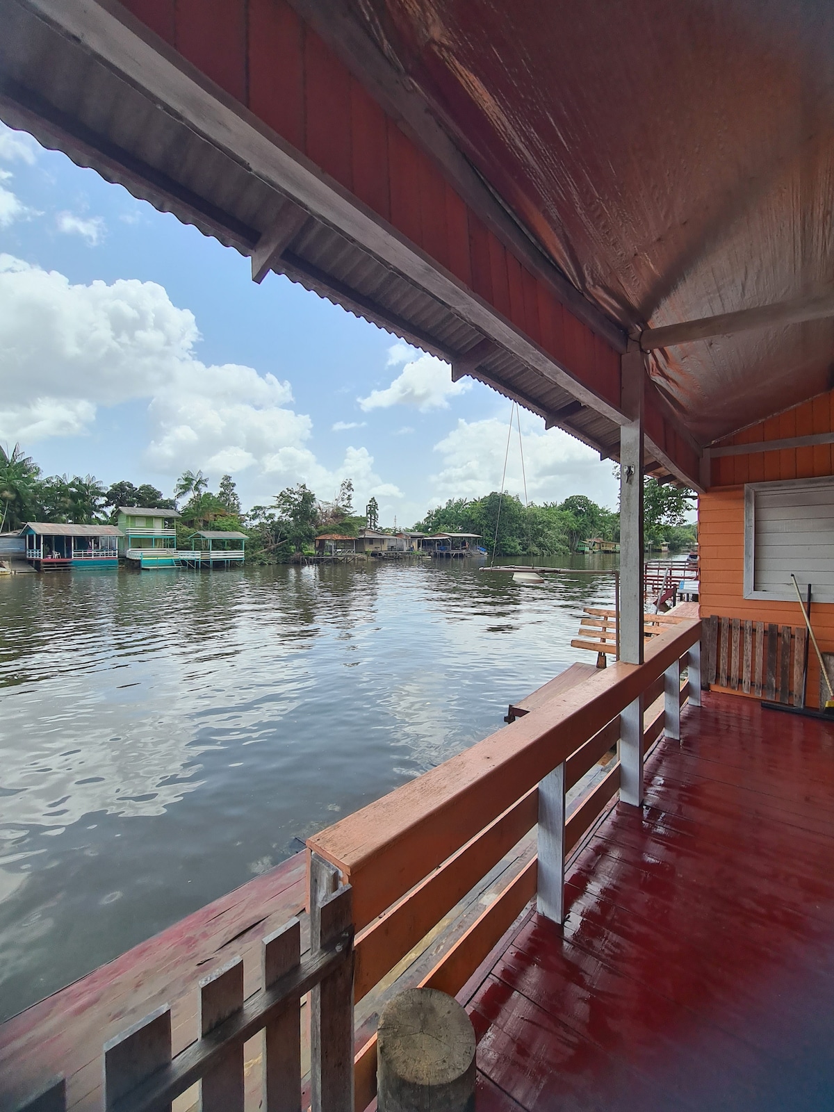 A covered outdoor deck overlooks a calm river, reflecting the blue sky and fluffy white clouds. Wooden railings frame the view, while various colorful houses and lush greenery are visible across the water, enhancing the serene ambiance of the surrounding environment.