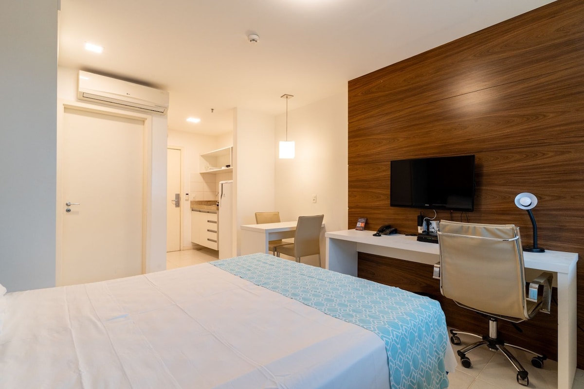 A modern room is displayed with a wooden accent wall behind a spacious bed covered in a light-colored duvet. A work desk with a chair and a television is visible. A kitchenette can be seen in the background, enhancing the functional aspect of the space.