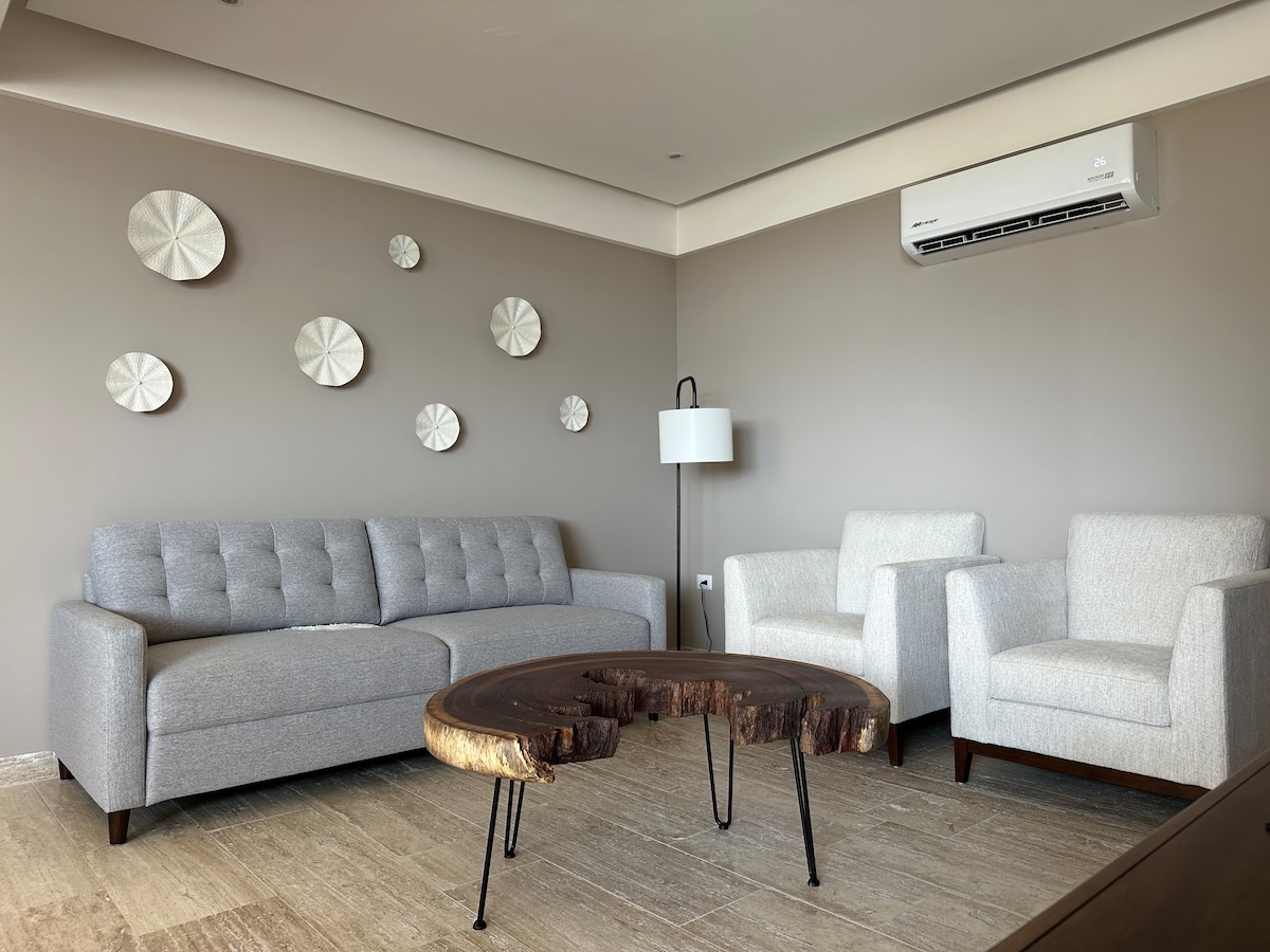 A modern living area is featured, showcasing a light gray sofa and two white armchairs. A unique wooden coffee table with an organic shape is positioned at the center. Decorative wall plates are arranged above the seating, complemented by a standing lamp in the corner.