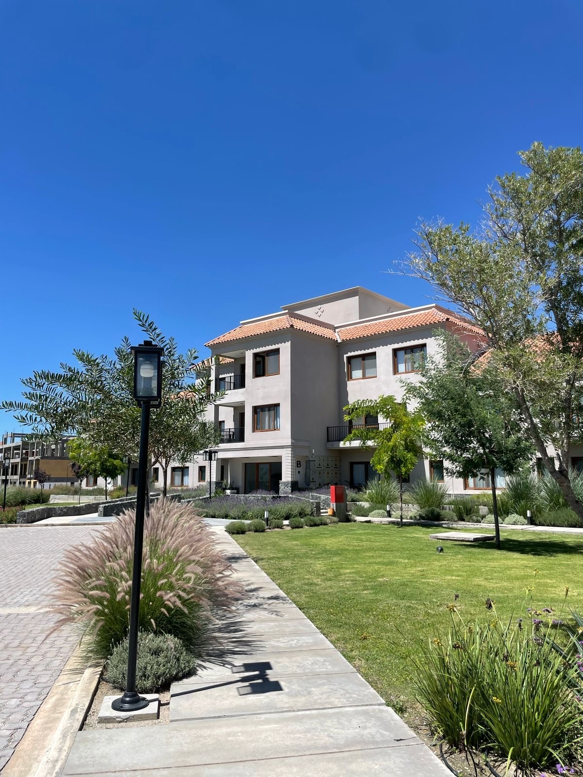 The image depicts a modern building within a well-kept complex, featuring a light-colored facade complemented by a terracotta-tiled roof. Surrounding the structure are landscaped areas with greenery, shrubs, and ornamental grasses, under a clear blue sky.