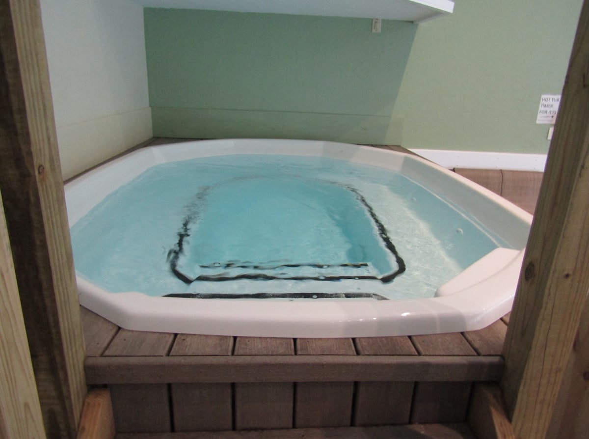 An indoor hot tub is set within a light green wall space, demonstrating a clean design. Water fills the tub, revealing a smooth bottom and surrounding wooden decking that enhances the serene environment. A small sign is visible nearby, adding to the space's functionality.