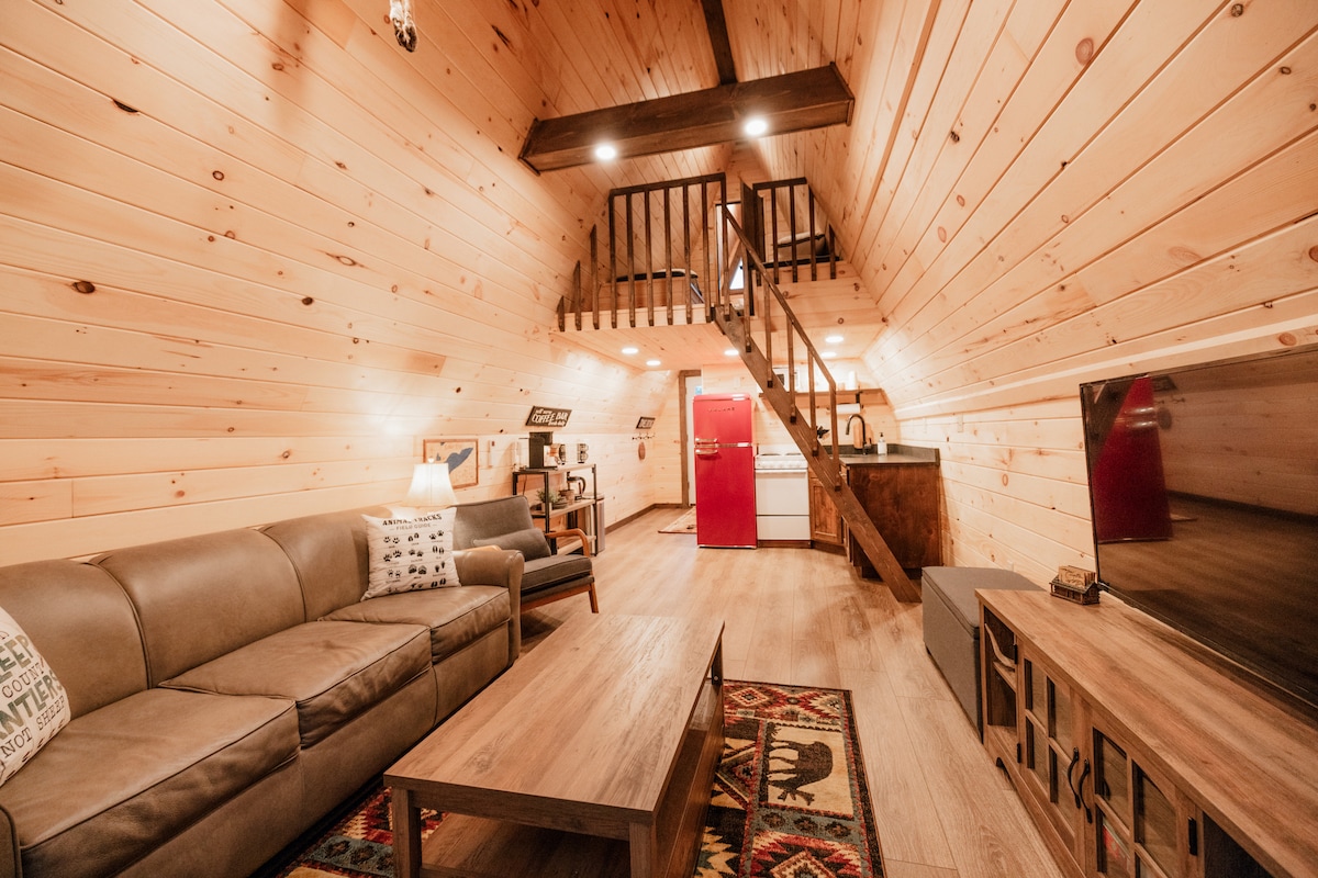 The living area features natural wooden walls and a vaulted ceiling, with a comfortable sofa and a coffee table positioned on a patterned rug. A staircase with ladder-style steps leads to the loft, while a red refrigerator is visible in the kitchen area.