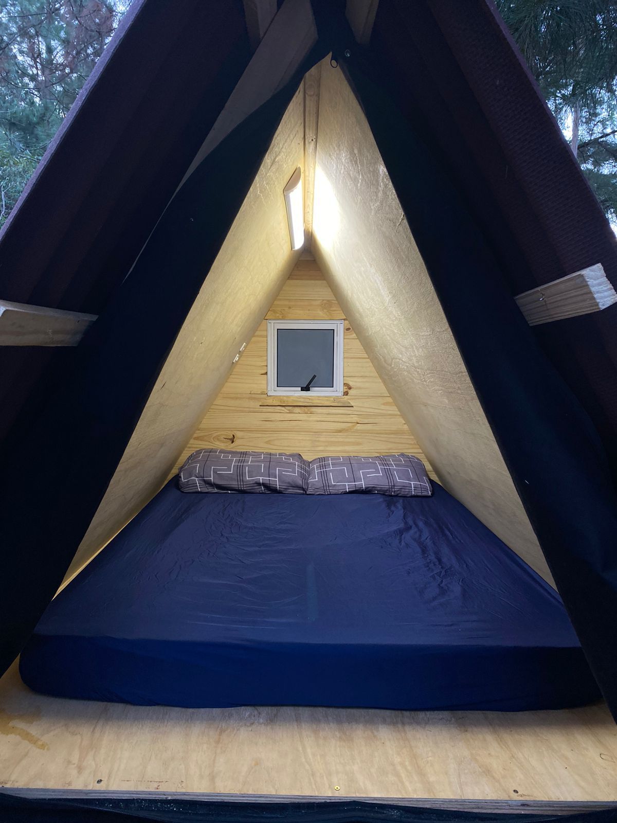 The interior of a wooden cabin is presented, featuring a spacious bed with a dark blue mattress and two gray pillows. A small window is visible on the opposing wall, allowing natural light to enter the cabin.