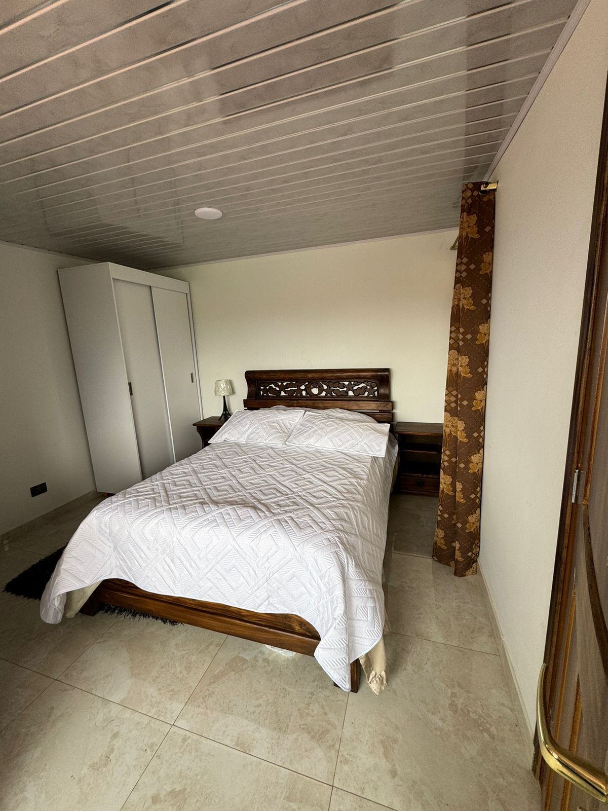 A spacious bedroom is presented, featuring a double bed with a textured white blanket. A wooden nightstand stands beside the bed, while a closet is visible against the wall. Soft lighting and a neutral color palette create a calming ambiance.