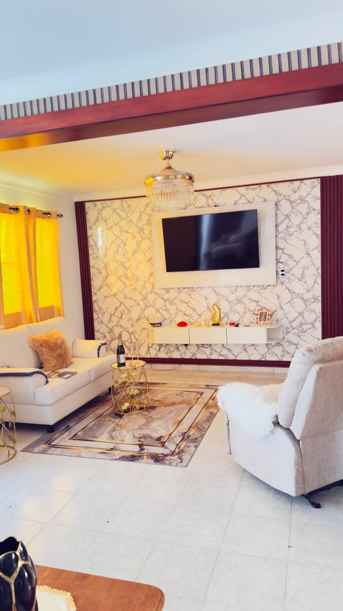 A modern living area is featured with a marble accent wall and a mounted television. A cozy white sofa and an armchair are positioned around a decorative area rug. Natural light is filtered through yellow curtains, enhancing the space's bright ambiance. A small coffee table adds functionality.