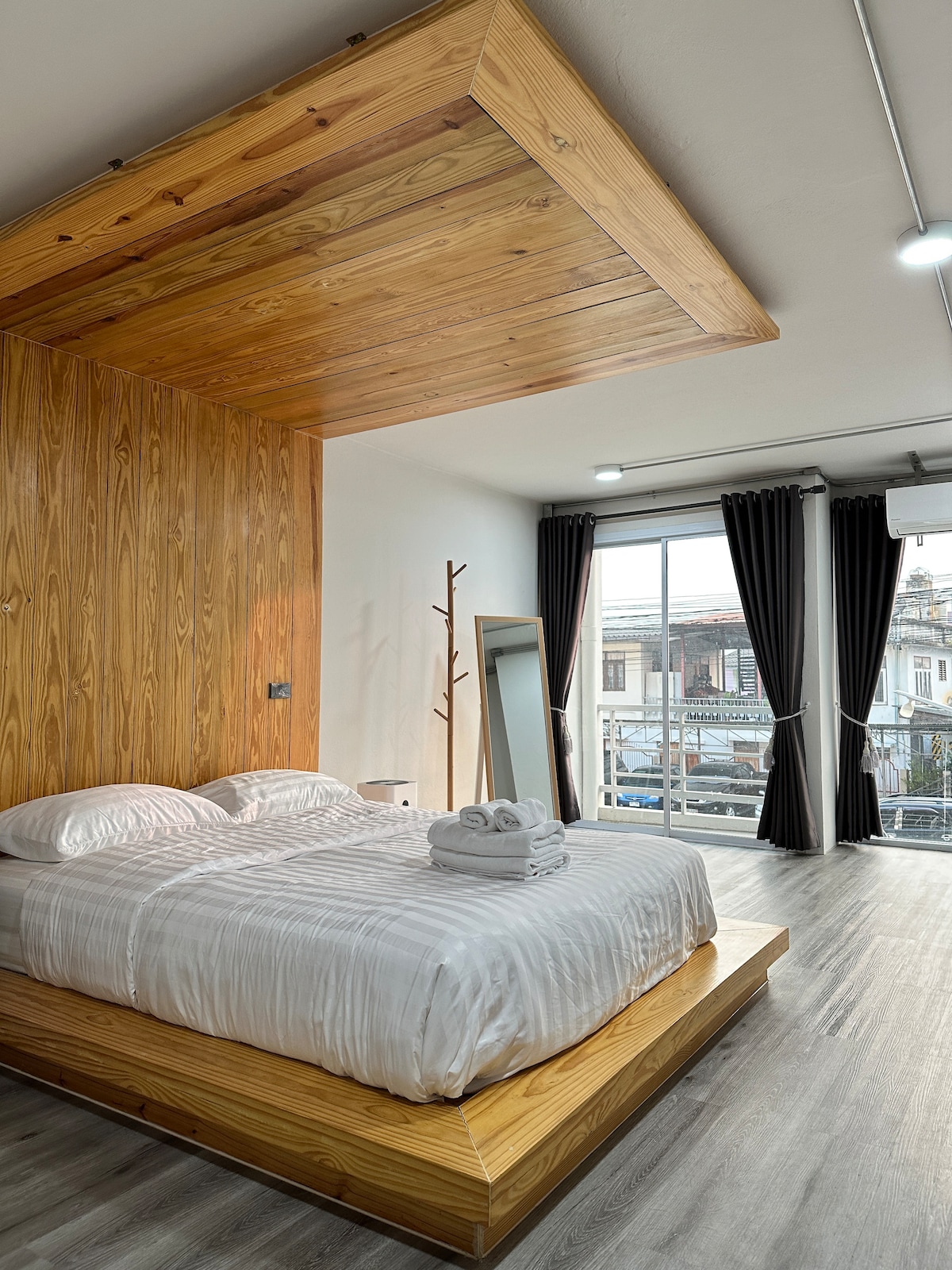 A wooden accent wall creates a warm backdrop for a spacious bed layered with soft linens. Natural light fills the room through large windows, with sleek dark curtains providing contrast. A minimalist mirror and a slender coat rack add functionality to the inviting space.