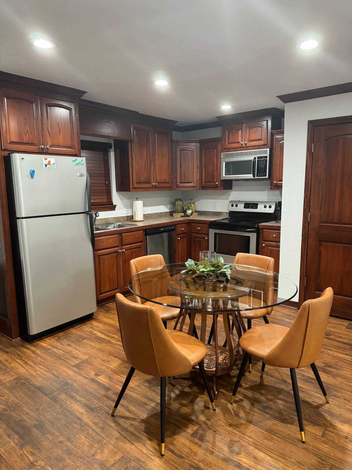 A well-equipped kitchen features rich wooden cabinetry and modern appliances. A round glass dining table is surrounded by four brown upholstered chairs. Bright lighting illuminates the space, highlighting the stainless steel refrigerator and stove. The floor is adorned with warm wooden tones.