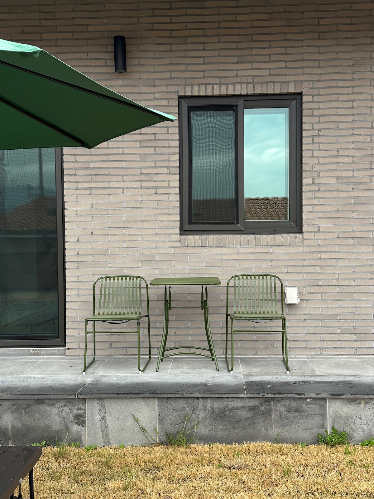 A small outdoor seating area is displayed with two green metal chairs and a matching round table. The setting is positioned next to a light-colored brick wall, featuring a window that allows natural light to enter the space.
