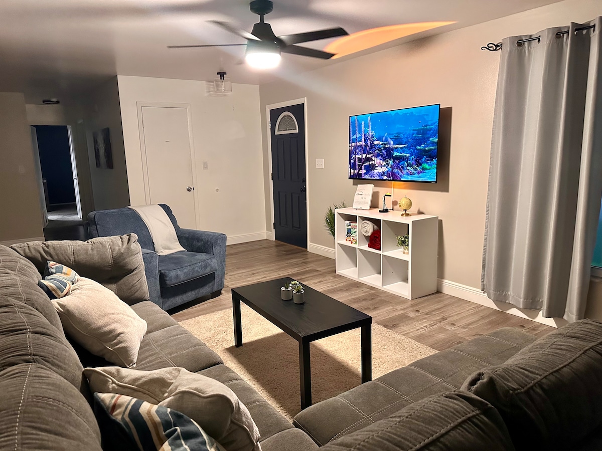A living area features a spacious gray sectional sofa, complemented by a cozy blue chair. A black coffee table occupies the center, while a television mounted on the wall displays an underwater scene. Soft lighting is provided by a ceiling fan and nearby window treatments.