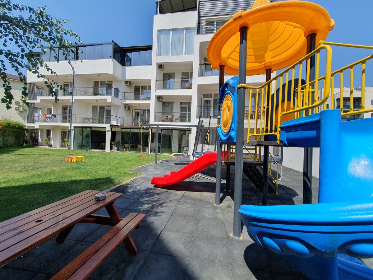 A spacious courtyard is featured, showcasing a colorful playground equipped with slides and climbing structures. Nearby, a wooden picnic table is positioned on a flat surface, surrounded by well-maintained grass. The backdrop includes a modern building with multiple balconies and large windows.