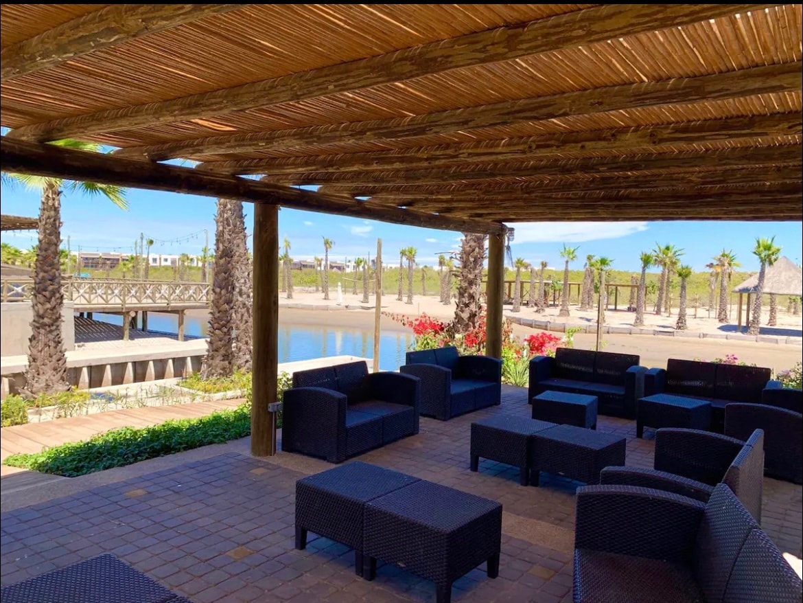 An outdoor seating area is sheltered by a wooden pergola, featuring dark wicker couches arranged around low tables. Lush greenery and blooming flowers frame the space, with a view of a serene waterway and palm trees in the background, contributing to a relaxed ambiance.