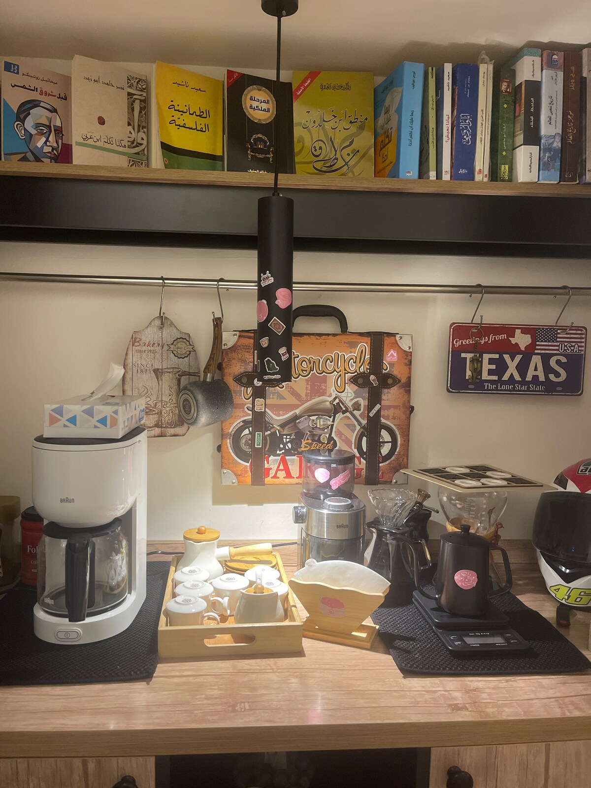 A well-equipped coffee station is presented, featuring a variety of brewing equipment, including a coffee maker and pour-over setup. A selection of mugs is organized on a tray, surrounded by fresh coffee beans. A collection of books and decorative signage adorns the shelf above.