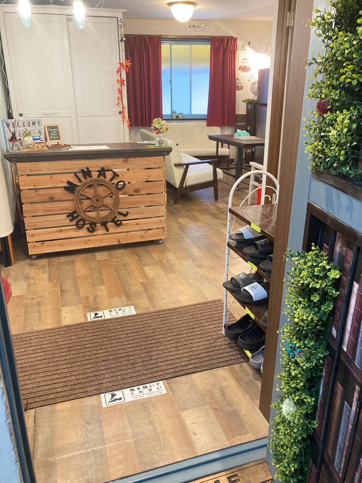 An entrance view into the hostel lobby is visible, featuring a welcoming reception desk made of wood. Shoes neatly line the floor, and a cozy seating area with a table is seen in the background. The space is adorned with warm lighting and decorative plants.