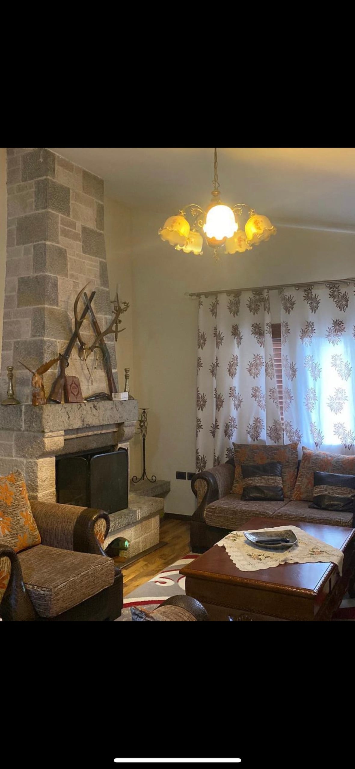 A cozy living room features a stone fireplace adorned with decorative antlers. A wooden table sits at the center, surrounded by upholstered chairs with patterned fabric. Light filters through floral curtains, creating a warm atmosphere in the space.