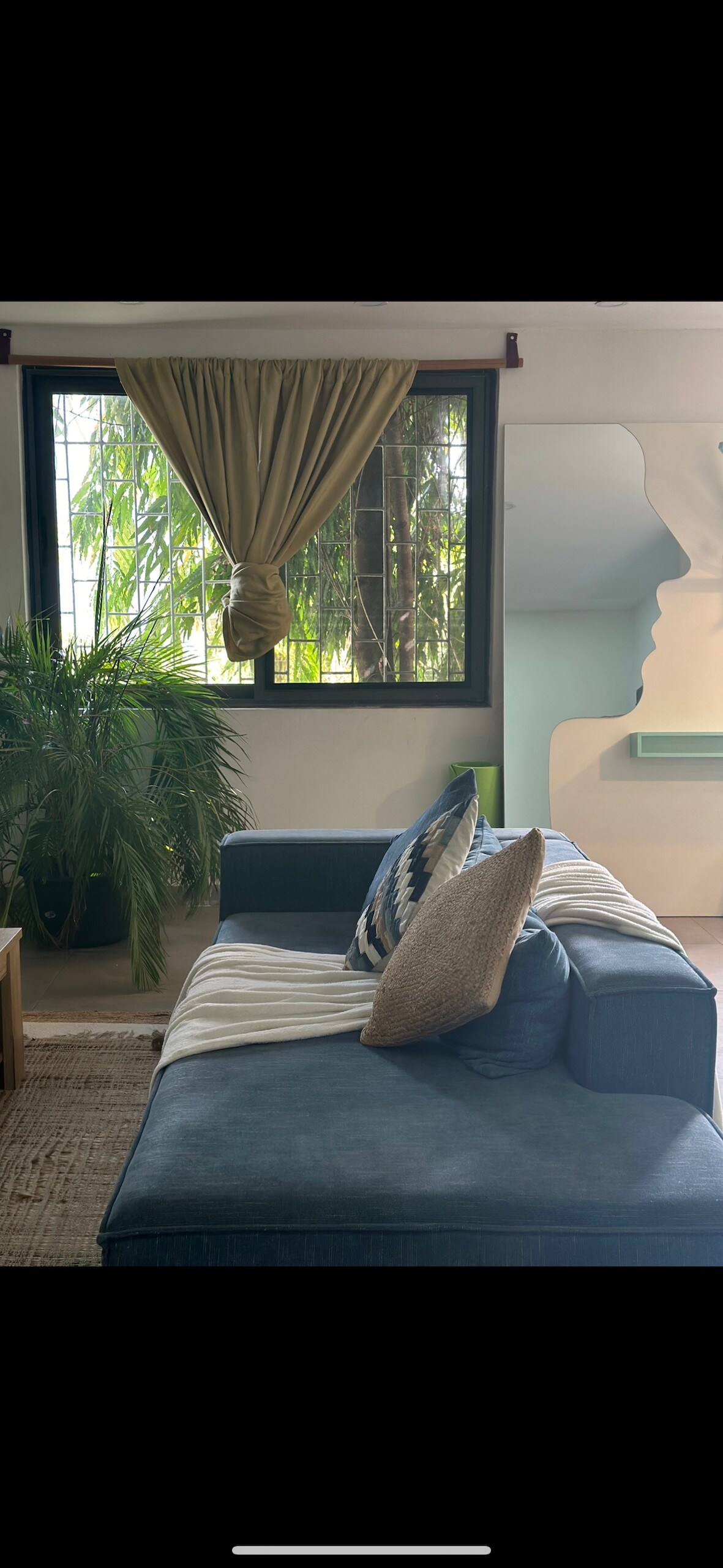 A comfortable blue sofa is positioned against a window, with sunlight filtering through sheer curtains. Lush greenery is visible outside, and decorative pillows add a touch of texture. A unique visage-shaped wall feature contributes to the room's artistic ambiance.