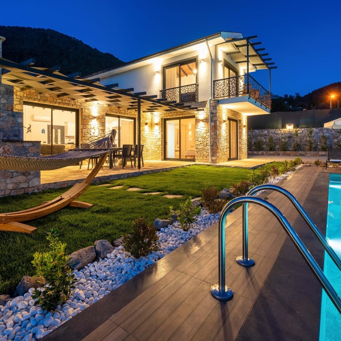 A two-story villa is showcased at dusk, featuring a stone exterior with modern design elements. A hammock is situated in the garden, along with a dining table and chairs. The inviting pool is bordered by lighting and lush green grass, enhancing the outdoor living space.