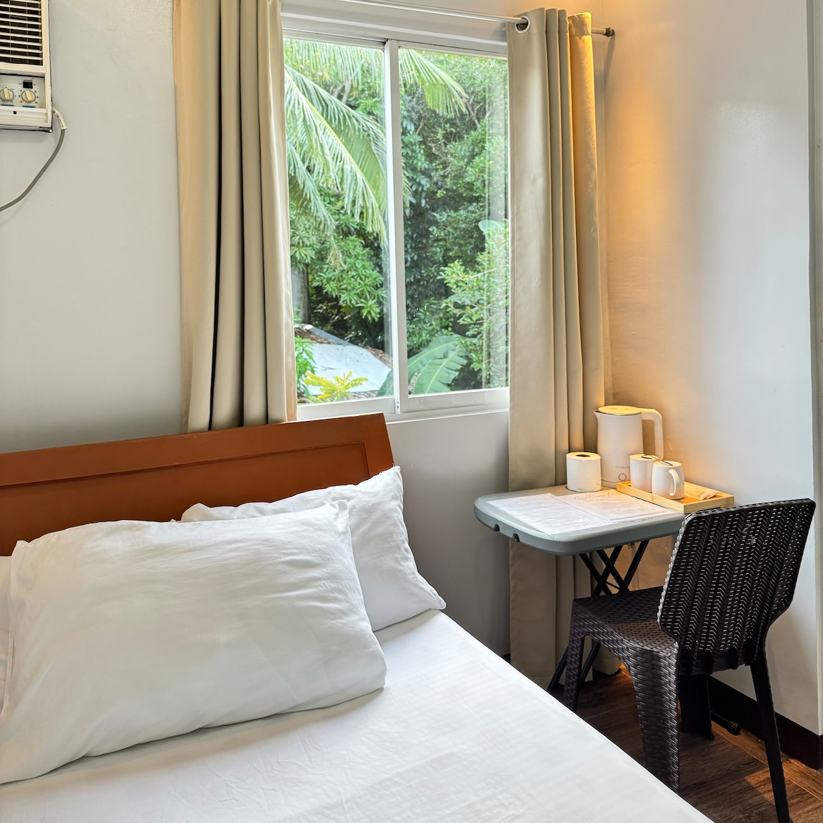 A cozy sleeping area features a double bed with simple bedding. Natural light enters through a window adorned with light curtains, illuminating a small desk with a chair beside it. Lush greenery is visible outside, contributing to a serene atmosphere.
