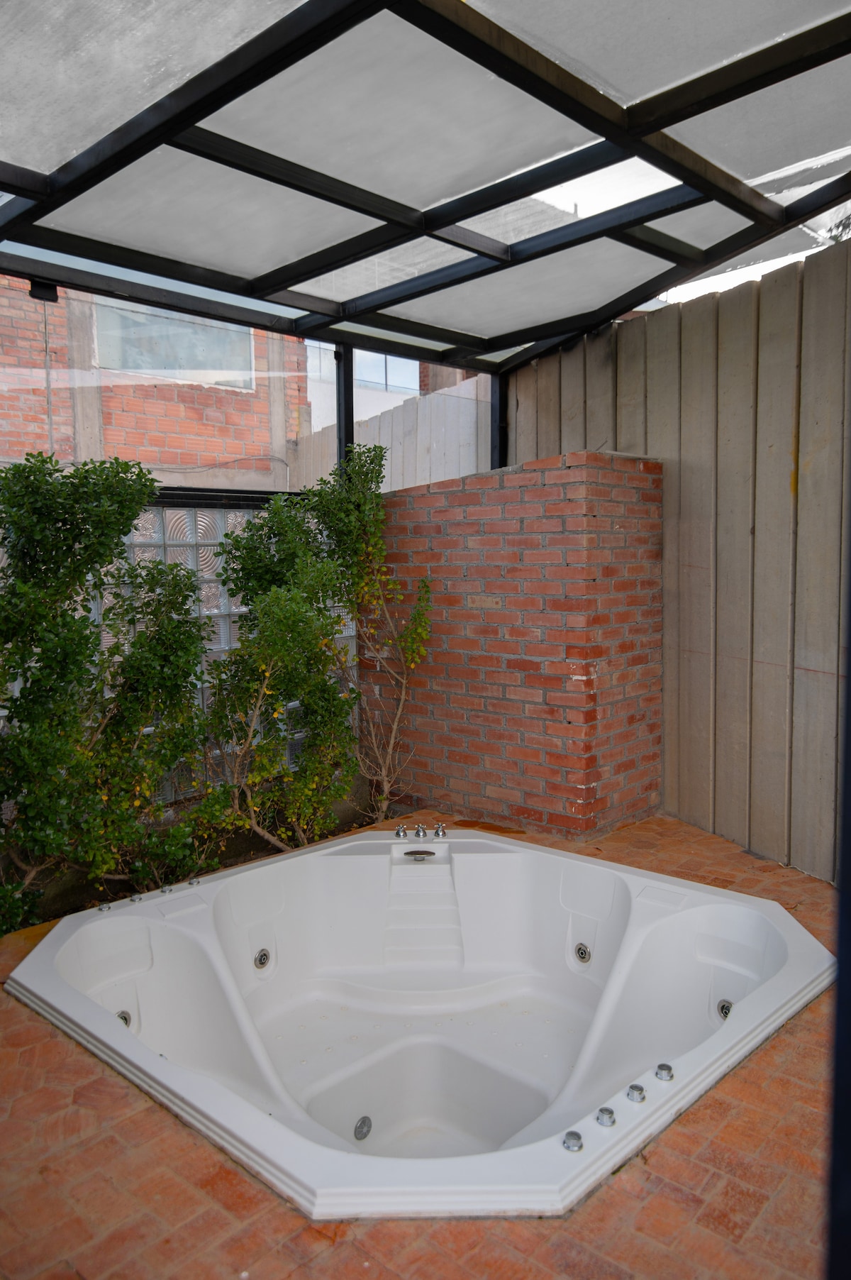 An outdoor jacuzzi is situated under a covered structure, surrounded by greenery and a brick wall. The space is designed for relaxation and privacy, with ample room for comfort.