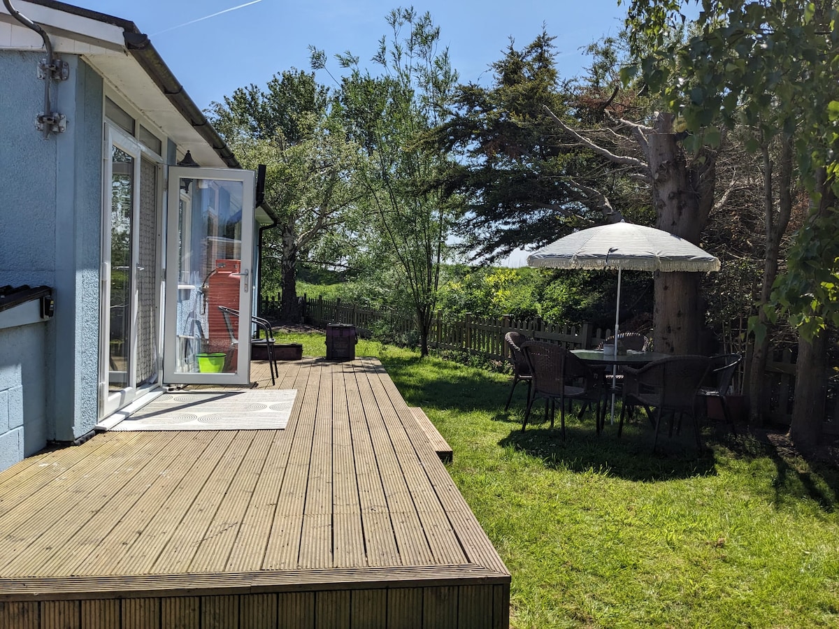 A wooden deck extends from the chalet, featuring a table set with four chairs under a large umbrella. Lush green grass surrounds the outdoor area, with a variety of trees providing shade in the background.