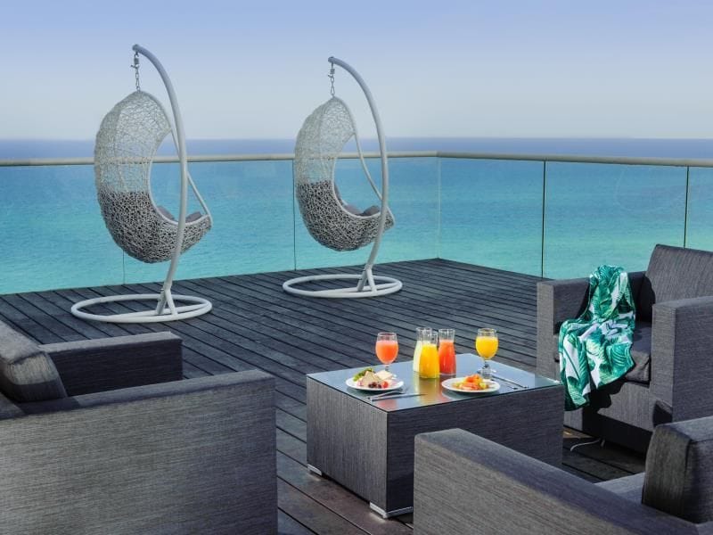 Two hanging chairs are positioned on a balcony with a glass railing, featuring sweeping views of the ocean. A coffee table holds colorful drinks and a plate of food. A green and white patterned blanket rests on a nearby chair, enhancing the outdoor lounge area.