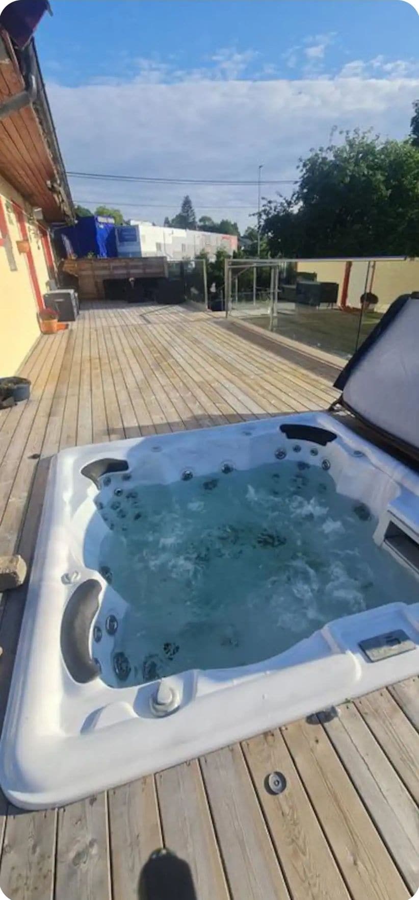 A hot tub is situated on a wooden deck, surrounded by a serene outdoor setting. The water is gently bubbling, and the area is enclosed with greenery, providing a private atmosphere. Sunlight filters across the deck, enhancing the inviting space for relaxation.