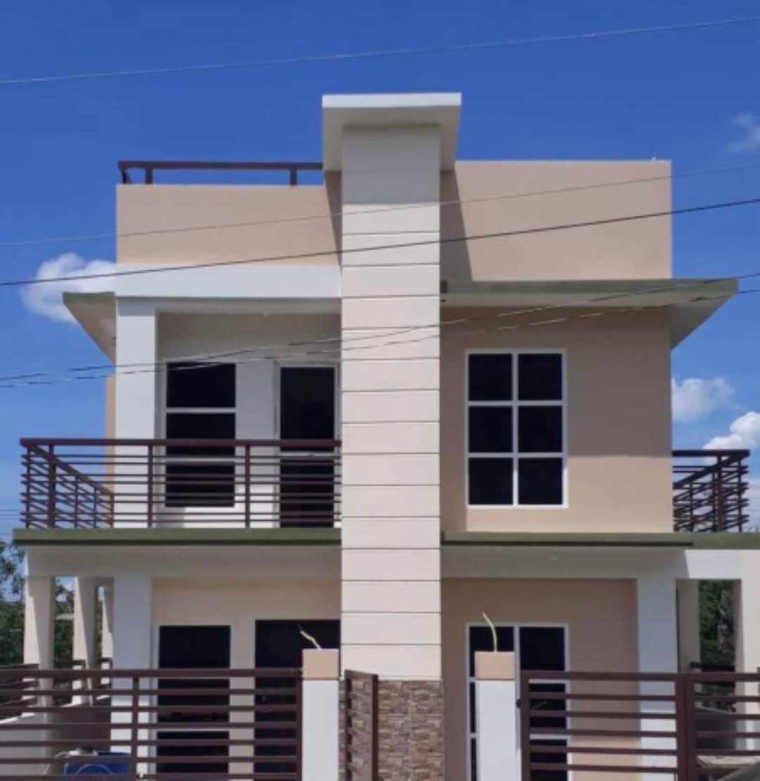The two-story building features a modern design with a combination of light beige and white exterior walls. Large windows with dark frames are visible on both levels, allowing for ample natural light. A balcony and a covered rooftop area add to the structure's appeal.