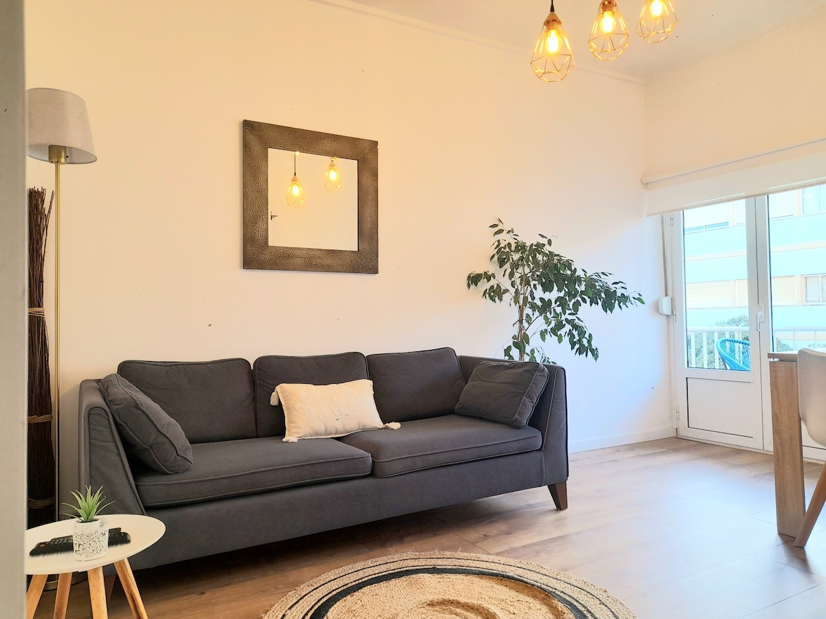 A comfortable living area features a gray couch with decorative pillows, positioned next to a small round table and a potted plant. A large window allows natural light to fill the room, complemented by hanging pendant lights and a framed mirror on the wall.