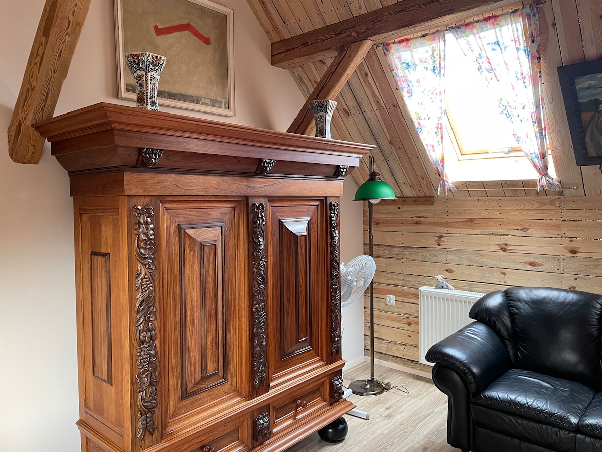 A carved wooden cabinet stands prominently in the room, showcasing intricate details. A green lamp adds a touch of color beside a black leather sofa. Natural light filters through a floral-draped window, illuminating the wooden walls and enhancing the inviting atmosphere.