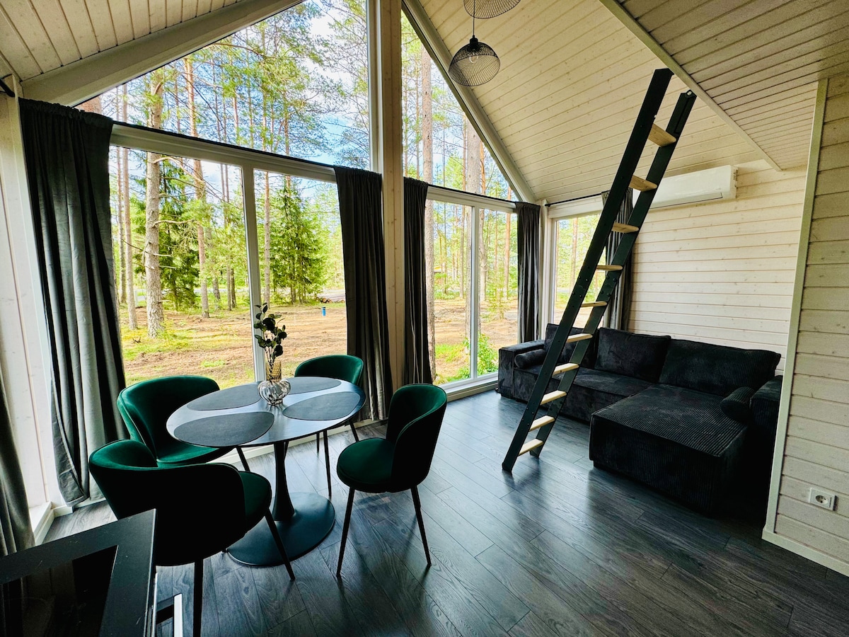 A bright living space is characterized by large windows that offer views of surrounding trees. A circular dining table with green chairs is positioned beside a dark couch, while a stylish ladder leads to the upper floor. Soft curtains frame the windows, allowing natural light to enter.