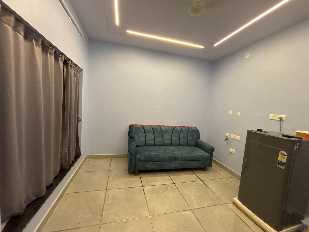 A small living area features a plush blue couch positioned near a large window, partially obscured by dark curtains. A stainless steel refrigerator stands nearby, complementing the modern aesthetic. The walls are painted a soft blue, and the floor is tiled in light gray.