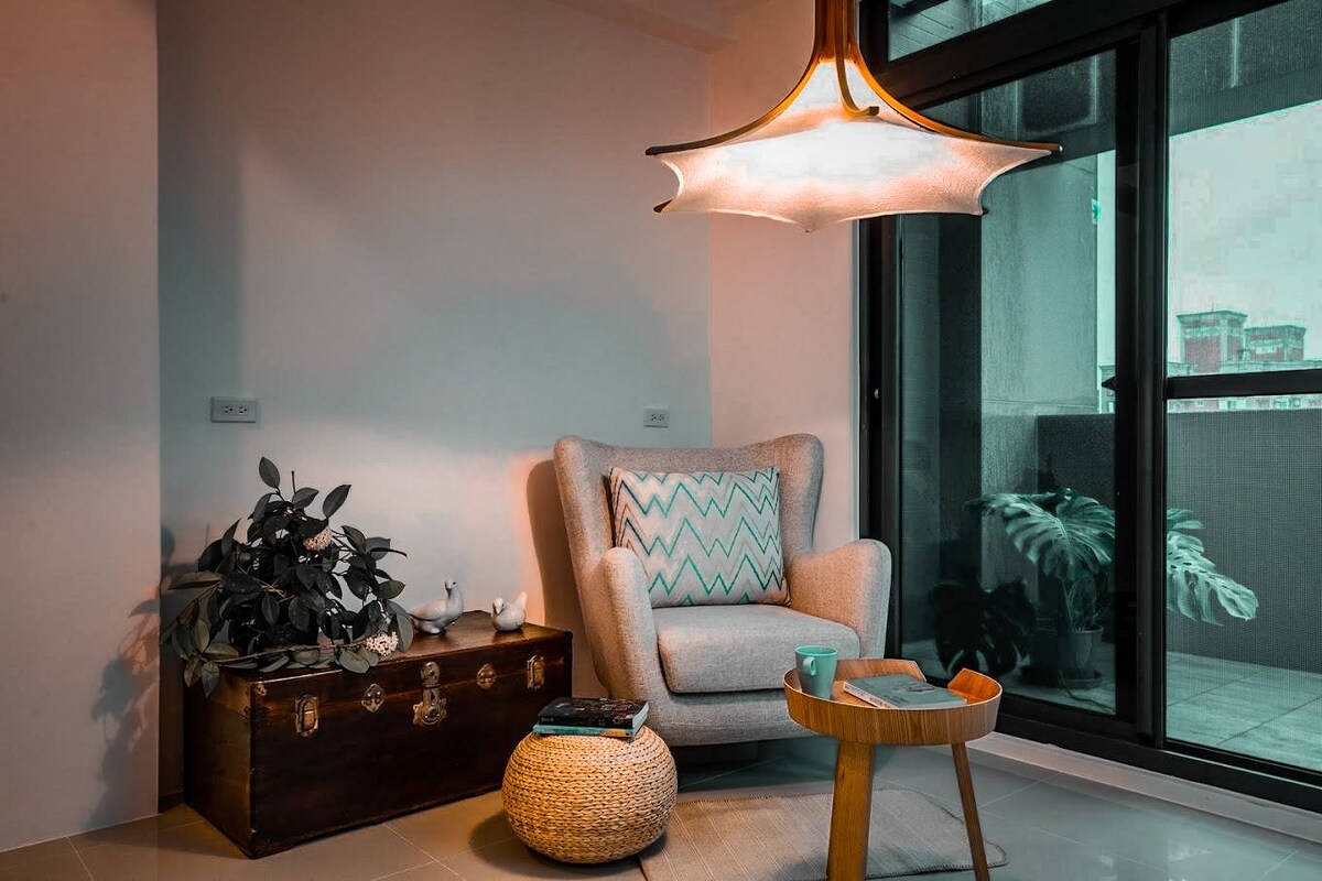 A cozy seating area is highlighted by a soft armchair adorned with a patterned cushion. A wooden side table supports a cup and decorative items, while a potted plant adds a touch of greenery. Large windows frame the view outside, allowing natural light to fill the space.