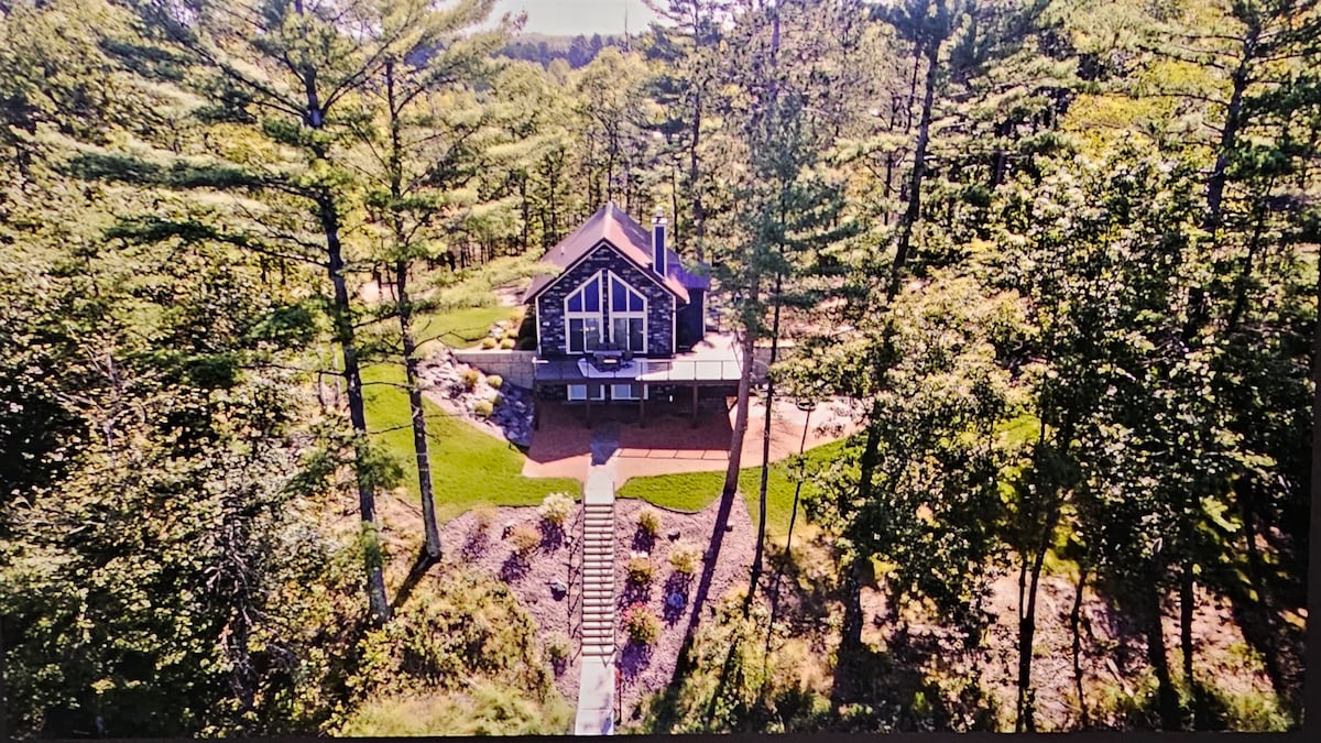 The cabin is surrounded by tall trees, showcasing its natural setting. A spacious deck is visible at the front, leading to manicured landscaping and a pathway that guides towards the entrance. The structure features large windows, inviting ample natural light.