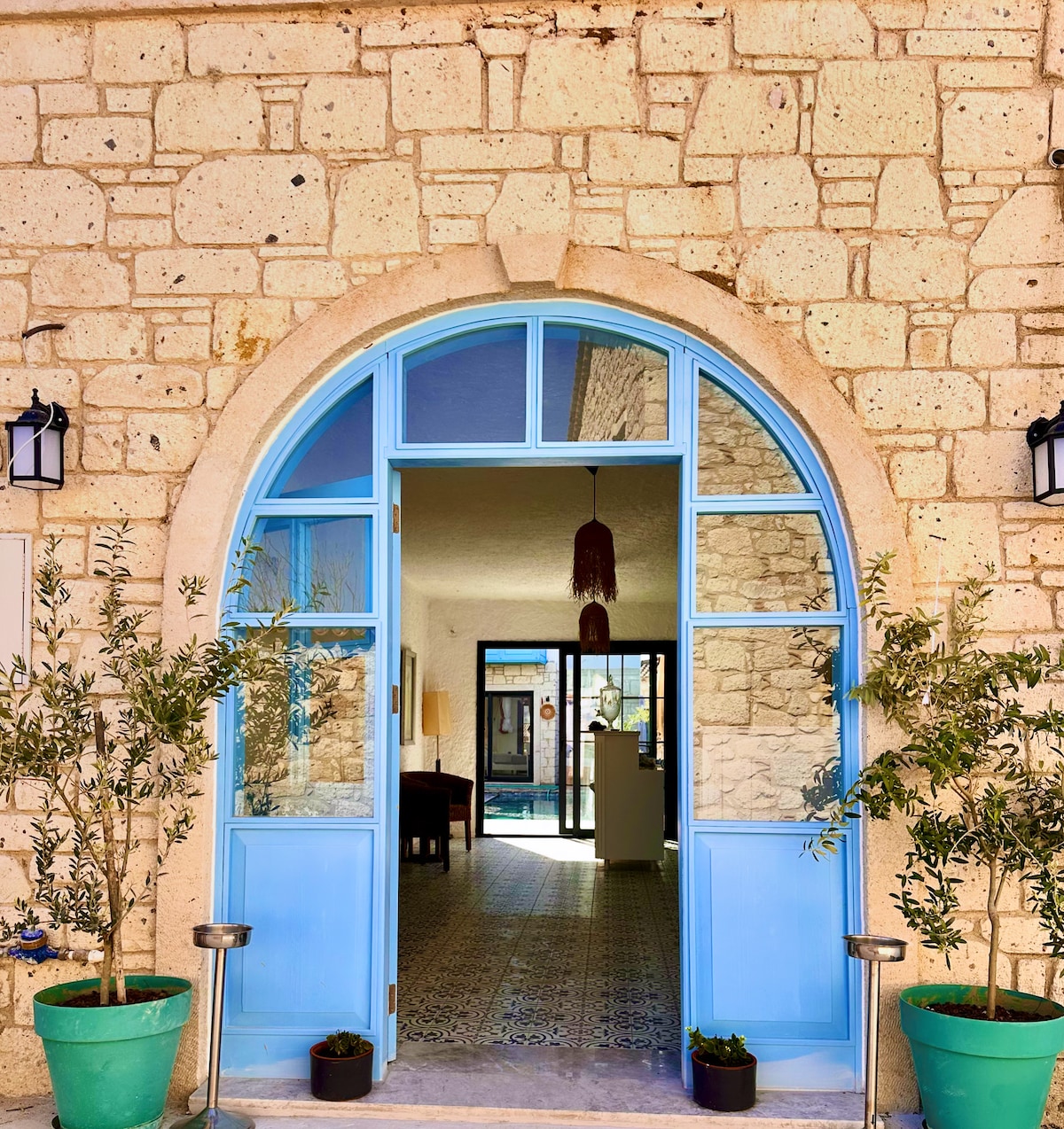 An inviting entrance showcases an arched doorway framed by light blue double doors. Potted plants are positioned on either side, enhancing the natural surroundings. The interior displays a glimpse of tiled flooring and a bright, open space beyond the entry, welcoming guests into the hotel.