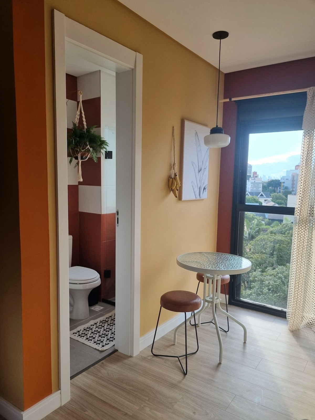 A small dining area features a round glass-topped table with two chairs, positioned near a large window. A modern light fixture hangs above the table, while a decorative wall art piece and a hanging plant add a touch of charm. Natural light illuminates the space.
