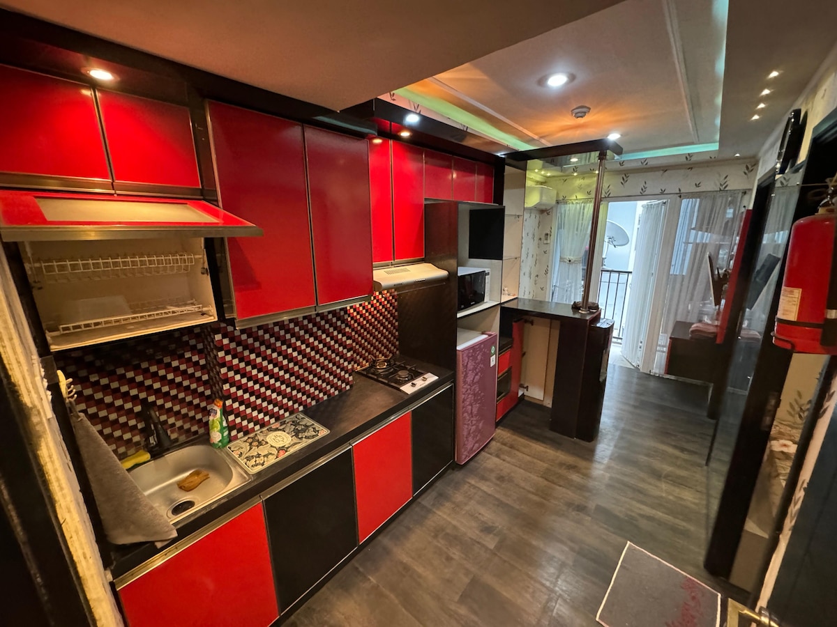 A modern kitchen is equipped with sleek red and black cabinetry. Features include a compact sink, a stove, and a microwave. A refrigerator is positioned nearby, with light reflecting off the glossy surfaces. Natural light enters from a nearby window.