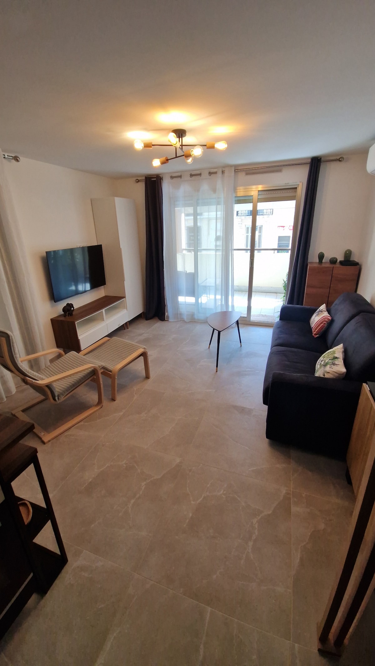 A comfortable living area features a dark sofa facing a wall-mounted television and a light wood coffee table. Natural light enters through sliding glass doors, which open to an outdoor space. Neutral-colored flooring complements the light walls and curtains.