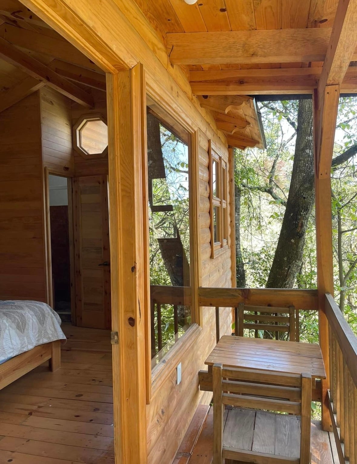 A wooden balcony features a small table and two chairs, surrounded by trees. Large windows in the adjacent room allow natural light to fill the wooden interior, enhancing the connection to the serene outdoor environment.