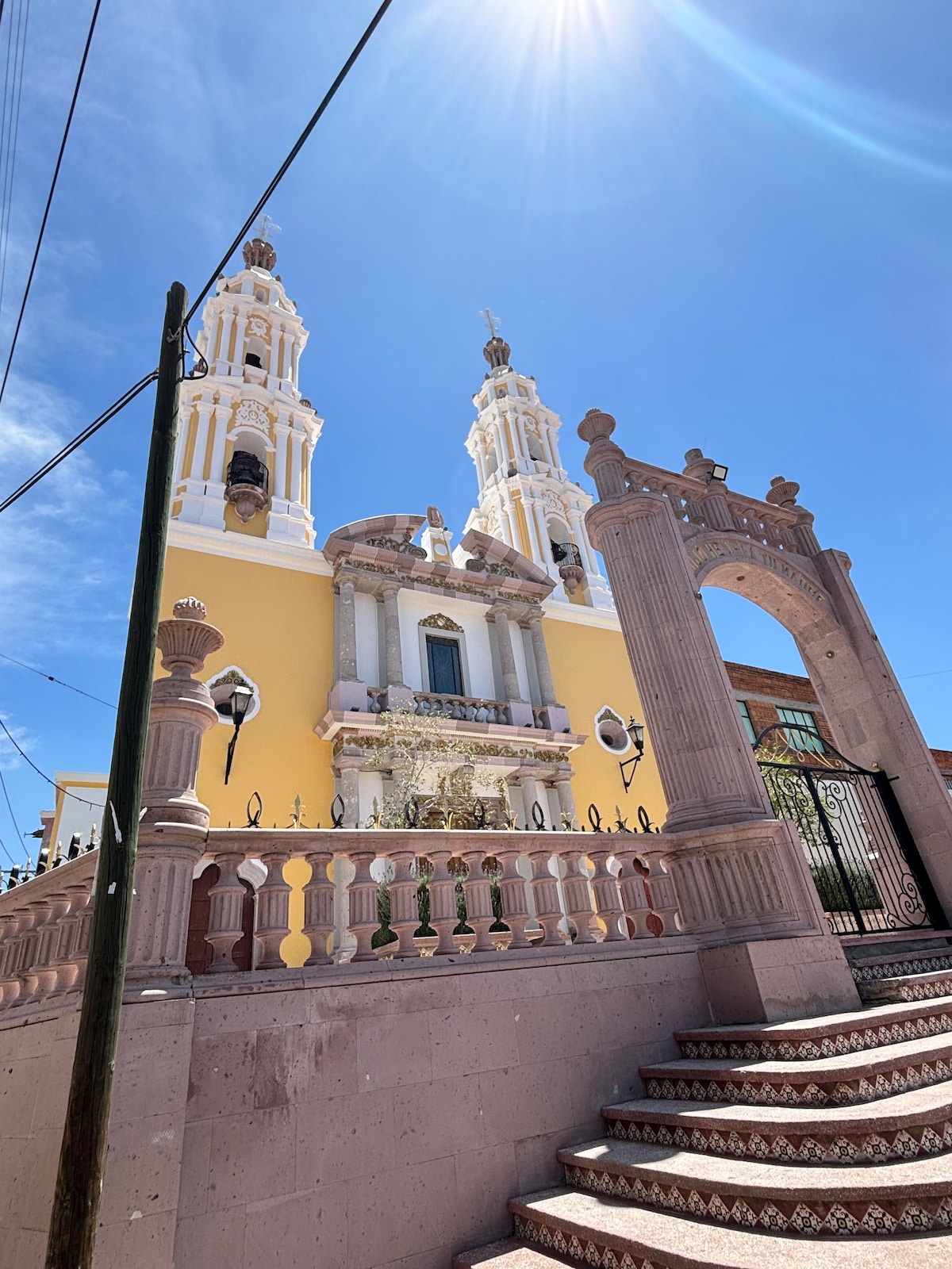 A striking church features two tall towers topped with ornate sculptures, showcasing a prominent façade painted in vibrant yellow. An intricately designed gate leads up to the entrance, flanked by decorative columns and balustrades, set against a clear blue sky.