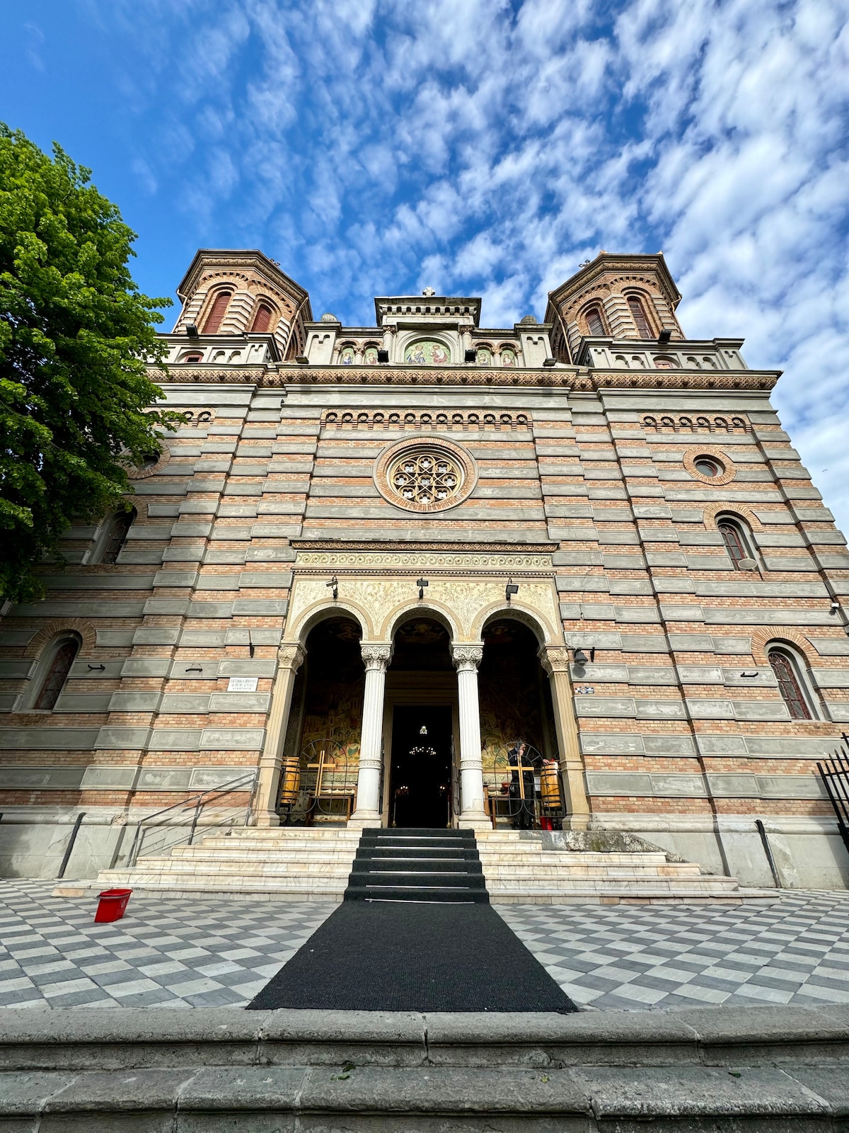 A large stone church features a facade with alternating light and dark stripes. The entrance is adorned with a carved archway and two towers rise above. A checkered pattern covers the ground leading to the steps. Lush trees border the building under a partly cloudy sky.