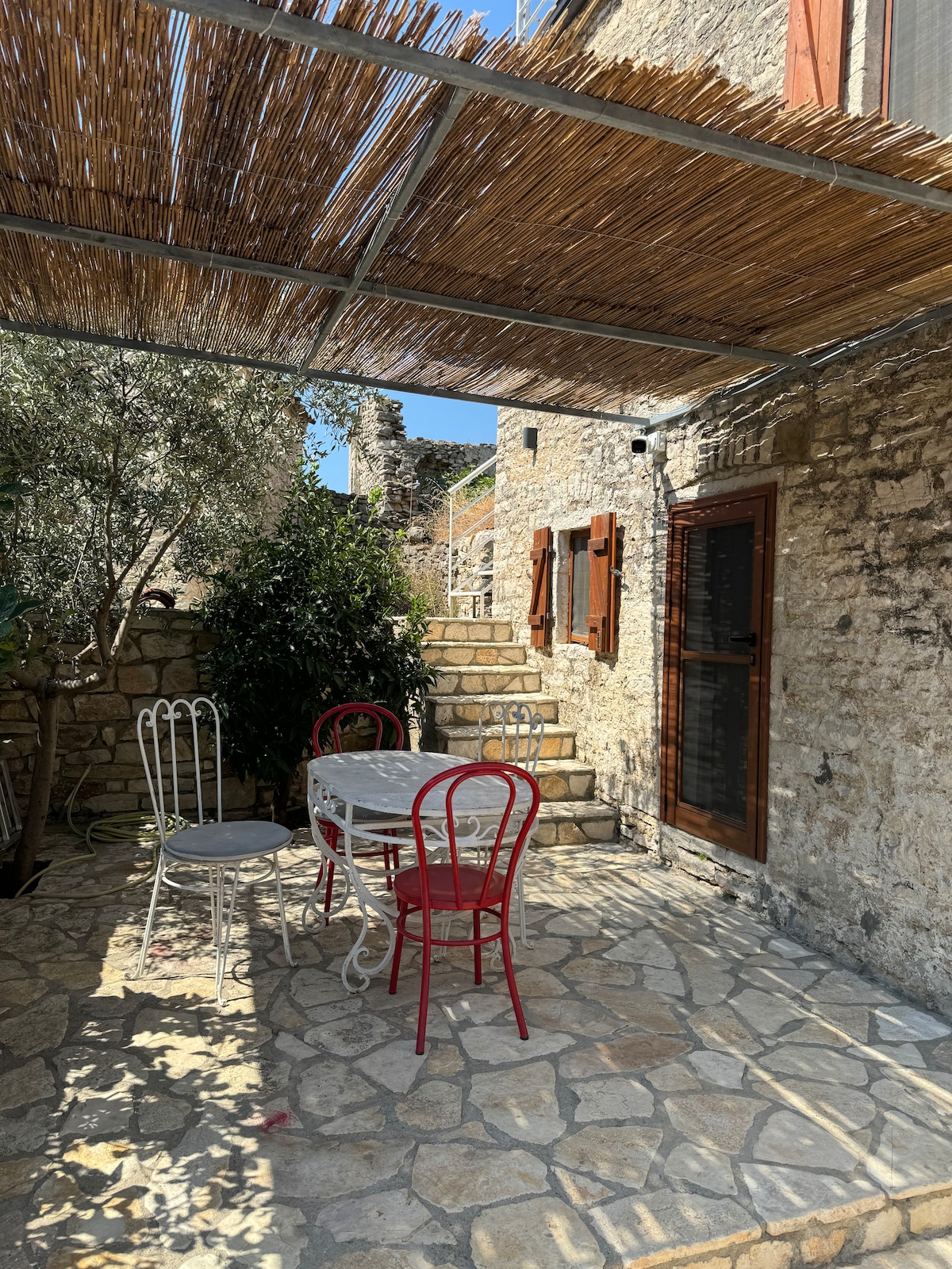 A charming outdoor area features a stone-paved patio with a table and four chairs, two white and two red. Natural shade is provided by a bamboo awning, and stone walls surround the space, complemented by greenery and steps leading to an elevated section.