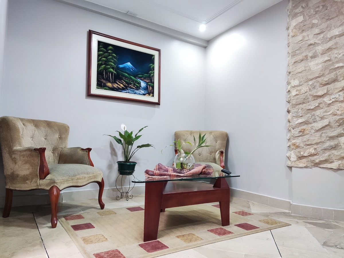 A corner of the living area is presented, featuring two upholstered chairs flanking a glass coffee table. A small plant and decorative items rest on the table, while an artwork depicting a mountain landscape adorns the wall above. Natural light enhances the inviting space.